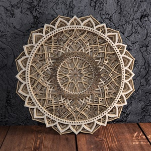 Könnte beinhalten: Ein detailliertes, kompliziertes Mandala-Design mit einem geschichteten 3D-Effekt. Das Mandala ist aus Holz und hat eine hellbraune Farbe. Es ist eine dekorative Wanddekoration oder ein Tischmittelpunkt.