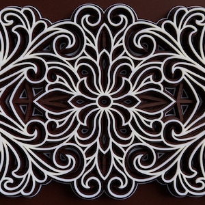 May include: A white paper cutout design with intricate floral patterns layered on top of each other. The design is symmetrical and has a brown background.