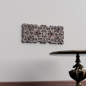 May include: A decorative wall hanging with a white intricate design on a brown background. The design features a floral pattern with swirls and curves.