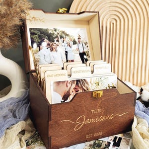 Puede incluir: Una caja de fotos de madera con tapa con bisagras y una inscripción personalizada que dice "The Jamesons Est 2025". La caja tiene divisores para organizar fotos e incluye una ranura para fotos en la tapa.
