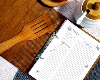 Puede incluir: Un archivador de recetas blanco con líneas negras y un diseño de abeja. El archivador está abierto en una página en blanco con las palabras "Receta", "Ingredientes" y "Instrucciones" impresas en ella. También se ve una espátula de madera, un azucarero blanco con tapa de madera y dos naranjas en la imagen.
