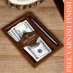 May include: A wooden tip jar with a slot for bills and a pen holder. The jar has a sign that says "Grill House" with five stars. The jar is sitting on a wooden surface.