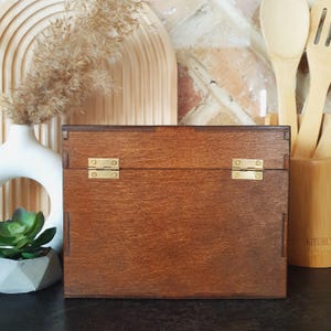 May include: A wooden box with a dark brown finish and brass hinges. The box is rectangular and has a simple, classic design. The background includes a white vase, decorative wooden elements, and kitchen utensils.