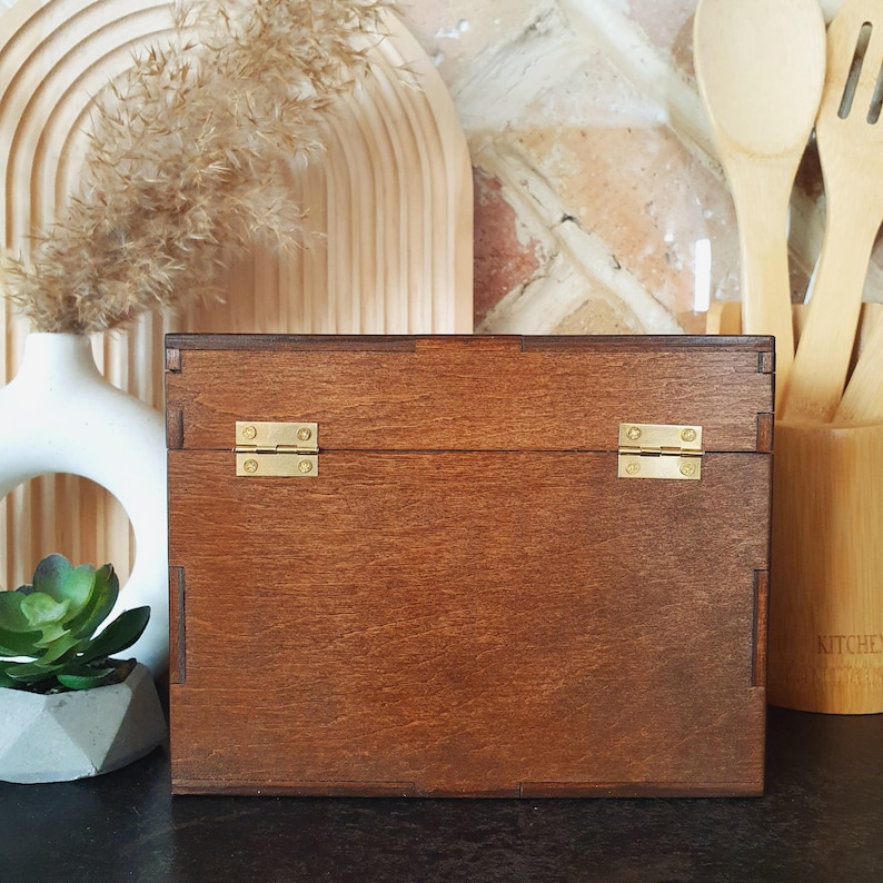 May include: A wooden box with a dark brown finish and brass hinges. The box is rectangular and has a simple, classic design. The background includes a white vase, decorative wooden elements, and kitchen utensils.