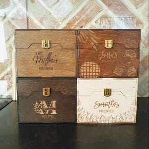 May include: Four wooden recipe boxes with gold latches. The boxes are in various shades of brown and white. Each box is engraved with a name and the word "RECIPES". One box has a waffle and cupcake design.