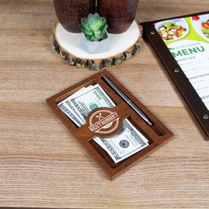 May include: A wooden tip jar with a slot for bills and a pen holder. The jar has the words "Restaurant" and a logo with crossed utensils on it. There are several US dollar bills inside the jar.