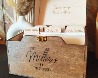 May include: Wooden recipe box with the text "The Miller's Recipes" engraved on the front. The box is open, revealing labeled dividers for "Pasta & Bakery," "Salads," and "Main Dishes." The inside lid has the quote "Every recipe tells a story."