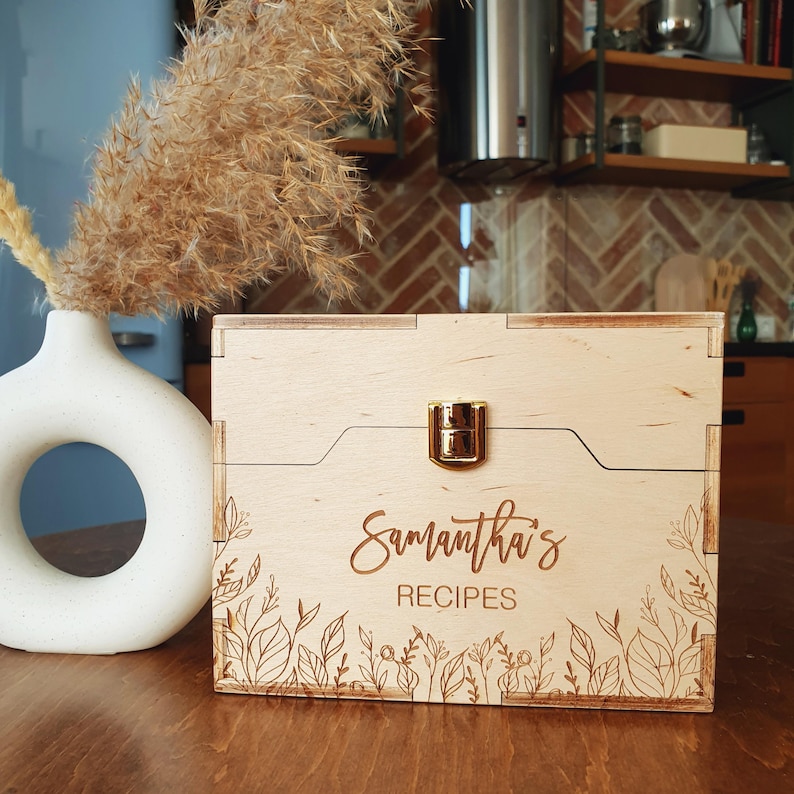 May include: A light-colored wooden recipe box with a gold-colored clasp. The box is engraved with the name "Samantha's" and the word "RECIPES" in a script font, along with floral designs. A white vase with dried plants is in the background.
