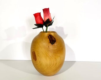 May include: A wooden vase with a natural wood grain finish. The vase is shaped like an egg and has a small opening at the top. Two red roses are in the vase.