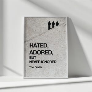 Puede incluir: Un cuadro enmarcado con el texto "Hated, Adored, But Never Ignored" en fuente negra. El cuadro tiene un fondo gris claro texturizado y un marco blanco. Las palabras "The Devils" están debajo del texto, con tres símbolos de flecha negros en la parte superior.