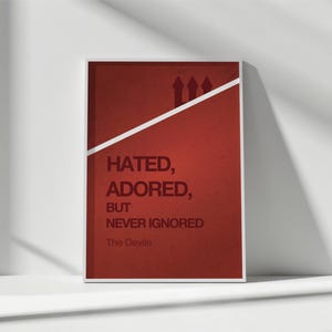May include: Framed red poster with a white border. The poster features the text "Hated, Adored, But Never Ignored" in a bold font. The words "The Devils" are at the bottom. A white diagonal line and three small silhouettes are at the top.