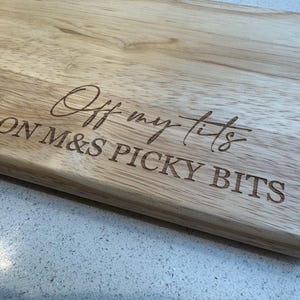 Könnte beinhalten: Rechteckiges Schneidebrett aus hellem Holz mit abgerundeten Ecken. Der Text "Off my tits ON M&S PICKY BITS" ist in Kursivschrift eingraviert. Die Holzmaserung ist sichtbar.