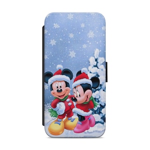 Puede incluir: Una funda de teléfono azul y blanca con una escena de invierno con Mickey Mouse y Minnie Mouse con gorros de Papá Noel. El fondo es un paisaje nevado con un pino.