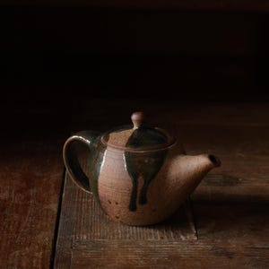 Può includere: Una teiera in ceramica marrone e verde con un coperchio. La teiera ha una glassa a goccia e si trova su una superficie di legno.