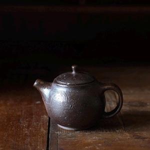 May include: A small, dark brown ceramic teapot with a lid and handle. The teapot has a rounded body and a spout for pouring. The surface has a textured appearance. The teapot is sitting on a wooden surface.