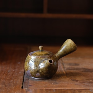 May include: A small, round, ceramic teapot with a side handle and a lid. The teapot is glazed in shades of olive green and brown, with a glossy finish. The spout is small and round. The teapot sits on a wooden surface.