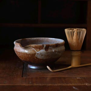 May include: A ceramic matcha bowl with a spout, featuring brown and white glaze, sits on a wooden surface. A bamboo whisk and scoop are also present, suggesting a tea ceremony. The bowl has a footed base.