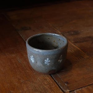 Może przedstawiać: Mały, ręcznie robiony ceramiczny kubek z nakrapianą, szaro-niebieską glazurą. Kubek posiada szereg białych kwiatowych wzorów wokół zewnętrznej strony. Kubek stoi na drewnianej powierzchni.