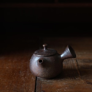 Op de afbeelding: Kleine, bruine keramische theepot met een afgeronde body, een korte tuit en een deksel met een kleine knop. De theepot staat op een houten oppervlak. De algehele esthetiek is rustiek en minimalistisch.