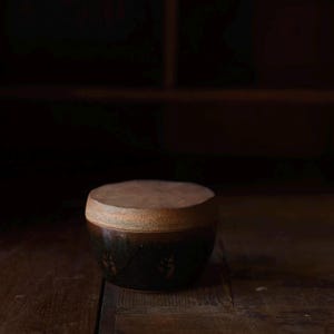 May include: A ceramic jar with a lid. The jar is dark brown with a textured surface and a lighter brown lid. The jar has a round shape and is sitting on a wooden surface. The jar has a decorative pattern on the side.