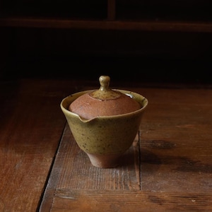 Może przedstawiać: Ceramiczna czarka do herbaty z pokrywką, posiadająca nakrapianą oliwkowozieloną glazurę na zewnątrz oraz podstawę w naturalnym kolorze terakoty. Czarka posiada mały dzióbek do nalewania z jednej strony oraz zaokrągloną pokrywkę z centralnym uchwytem.