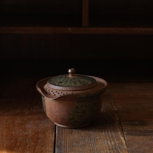 Puede incluir: Una tetera de cerámica moteada en marrón y verde oscuro con tapa a juego. La tetera cuenta con un colador integrado con pequeños orificios circulares cerca del pico y un cuerpo redondeado con un acabado terroso y texturizado.