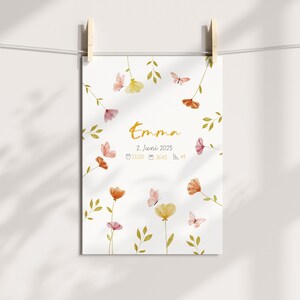 K&ouml;nnte beinhalten: Ein wei&szlig;er Druck mit dem Namen "Emma" in goldener Schrift, umgeben von Aquarellblumen und Schmetterlingen. Der Druck enth&auml;lt das Datum "2. Juni 2025" und weitere Details. Der Druck wird von Holzw&auml;scheklammern gehalten.