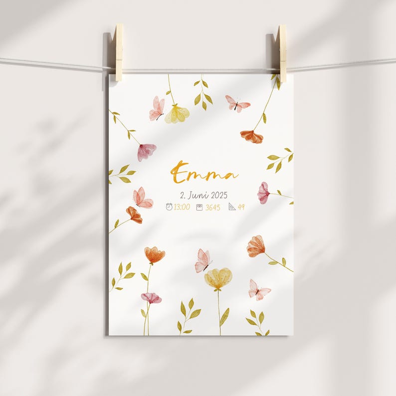 K&ouml;nnte beinhalten: Ein wei&szlig;er Druck mit dem Namen "Emma" in goldener Schrift, umgeben von Aquarellblumen und Schmetterlingen. Der Druck enth&auml;lt das Datum "2. Juni 2025" und weitere Details. Der Druck wird von Holzw&auml;scheklammern gehalten.