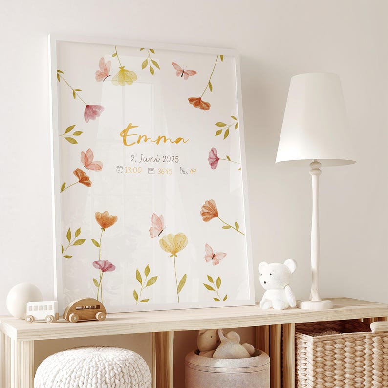 K&ouml;nnte beinhalten: Ein gerahmter Aquarelldruck mit dem Namen "Emma" in goldener Schrift, umgeben von floralen und Schmetterlingsillustrationen in zarten Rosa-, Orange- und Gelbt&ouml;nen. Der Druck enth&auml;lt Datum, Uhrzeit und Ma&szlig;e. Der Rahmen ist wei&szlig;.