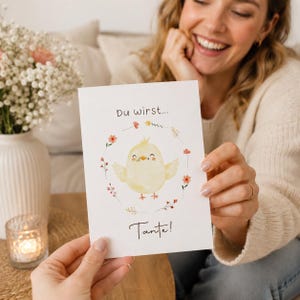 Könnte beinhalten: Eine weiße rechteckige Grußkarte mit einer Aquarellillustration eines gelben Kükens in der Mitte eines zarten Blumenkranzes. Der Text "Du wirst..." steht über dem Küken und das Wort "Tante!" ist darunter aufgedruckt.