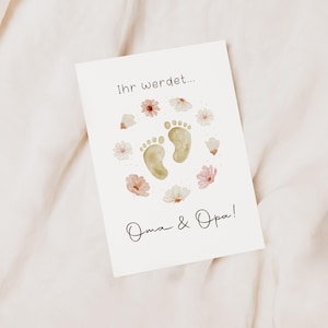 Könnte beinhalten: Eine weiße Karte mit dem Text "Ihr werdet..." und "Oma & Opa!" in schwarzer Schrift. Die Karte zeigt zwei hellbraune Babyfüße, umgeben von Aquarellblumen in Rosa und Weiß.