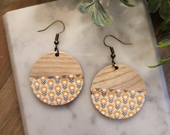 May include: A pair of wooden earrings with a white and yellow floral pattern. The earrings are round and have a gold-toned hook.