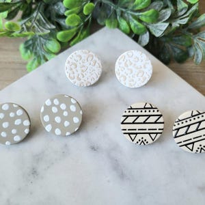 May include: A collection of six round stud earrings with various patterns. Two earrings have a leopard print design, two have white polka dots on a gray background, and two have black geometric patterns on a cream background.