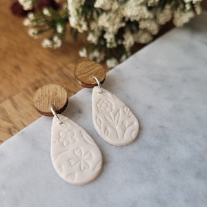 May include: A pair of white teardrop-shaped earrings with floral designs. The earrings are made of polymer clay and have wooden backs.