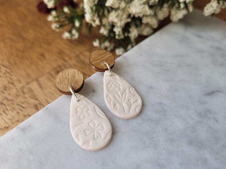 May include: A pair of white teardrop-shaped earrings with floral designs. The earrings are made of polymer clay and have wooden backs.