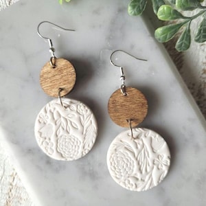 May include: A pair of dangle earrings with a white ceramic disc featuring an embossed floral design and a brown wooden disc. The earrings have silver hooks.