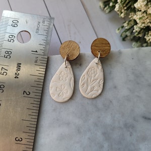 May include: A pair of white ceramic teardrop earrings with a floral design. The earrings are attached to wooden posts.