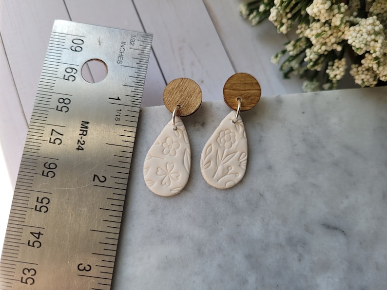 May include: A pair of white ceramic teardrop earrings with a floral design. The earrings are attached to wooden posts.