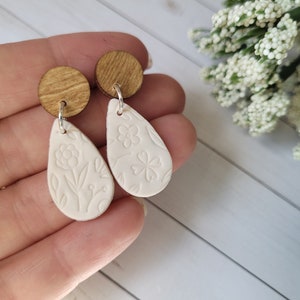 May include: A pair of white teardrop-shaped earrings with floral designs. The earrings are made of polymer clay and have wooden tops.