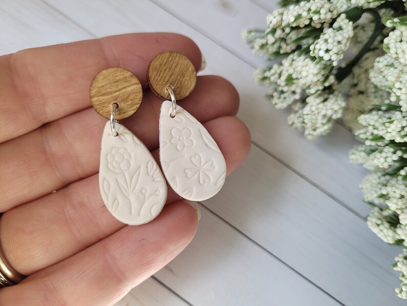 May include: A pair of white teardrop-shaped earrings with floral designs. The earrings are made of polymer clay and have wooden tops.