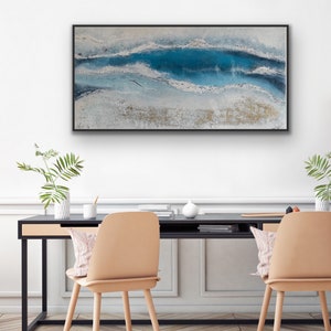 May include: Abstract ocean-themed painting in a black frame. The artwork features a textured surface with shades of blue, white, and gold, creating a sense of depth and movement. The painting is displayed above a black desk with two beige chairs.