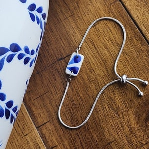 May include: A silver chain bracelet with a blue and white ceramic charm. The charm is rectangular and has a swirling pattern.