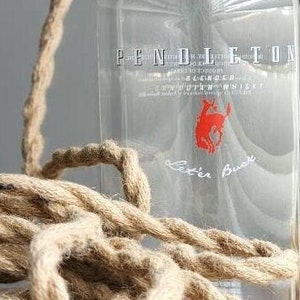 May include: A clear glass bottle pendant light fixture featuring the Pendleton blended scotch whisky label with a red buck logo. The lamp includes a black socket, a black power cord with a switch, and a thick, twisted tan jute rope cord cover.