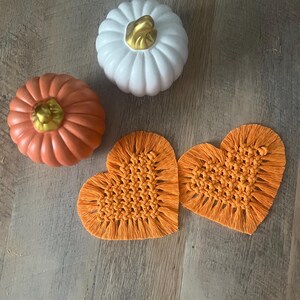 May include: Two heart-shaped macrame coasters made of orange cord with fringed edges, accompanied by one white pumpkin and one orange pumpkin, both featuring metallic gold stems. The items are arranged together on a flat surface.