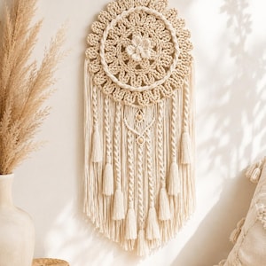 Puede incluir: Un tapiz de macramé color crema que presenta una parte superior circular tejida con un motivo floral central y un borde festoneado. Debajo del círculo, cuelgan múltiples cordones largos, acentuados con secciones de cuerda trenzada y borlas.