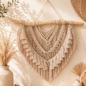 Puede incluir: Un tapiz de macramé beige con base de madera flotante. El intrincado diseño presenta patrones anudados y borlas escalonadas, creando una estética bohemia. La decoración de la pared está suspendida por un cordón de fibra natural.