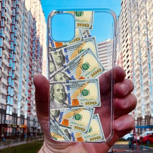 Puede incluir: Una funda transparente para teléfono con un diseño de billetes de dólar estadounidense. Los billetes están dispersos aleatoriamente y presentan el retrato de Benjamin Franklin y el texto "100" en la esquina superior derecha.