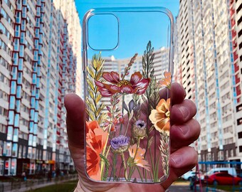 May include: A clear phone case with a floral design featuring red, yellow, and purple flowers. The case is held in a hand against a backdrop of apartment buildings.