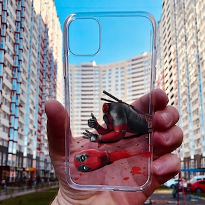 Puede incluir: Una funda transparente para teléfono con un gráfico de Deadpool tirado en el suelo con sangre salpicada a su alrededor. La funda se sostiene en una mano con un fondo borroso de edificios.
