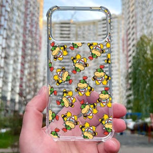 May include: Clear phone case featuring a repeating pattern of a cartoon character with a yellow body, green shell, and red hair. The case is held up against a blurred background of buildings.